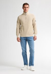 Beige ribbad stickad tröja med halvhög krage och dragkedja, i kombination med ljusblå jeans och vita sneakers. Har ett logotypaccenter på ärmen.