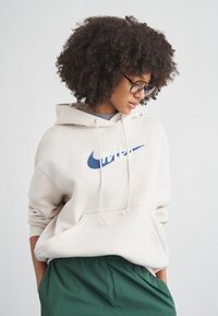 Sweatshirt bege com logótipo Nike Swoosh azul-marinho, cordões e bolso frontal; combinado com calças verdes texturizadas.