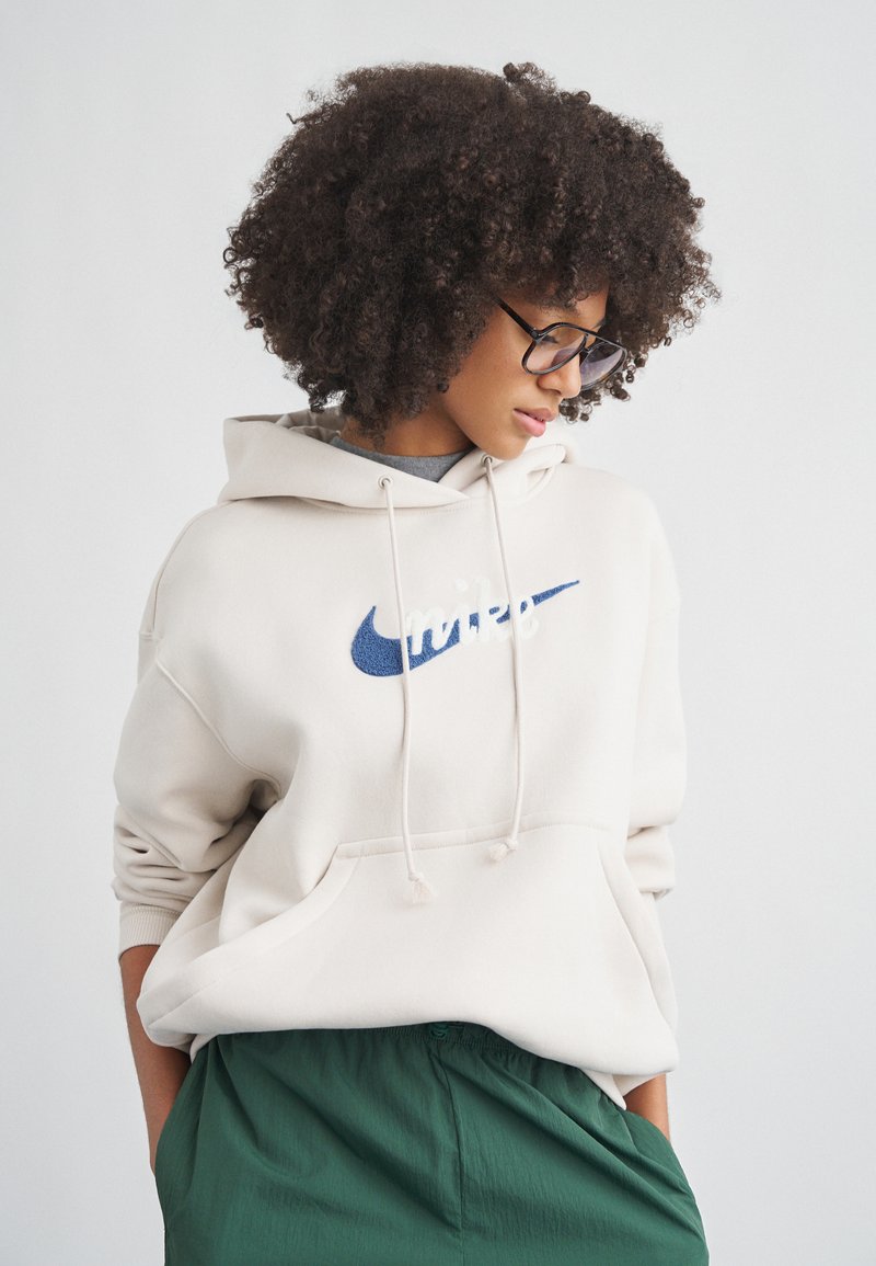 Sweatshirt bege com logótipo Nike Swoosh azul-marinho, cordões e bolso frontal; combinado com calças verdes texturizadas.