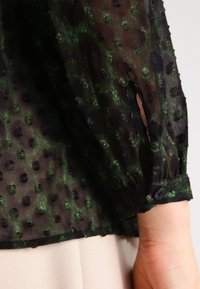 Blouse noire transparente avec des pois texturés verts et noirs, dotée de manches longues froncées et d'un détail de manchette boutonnée.