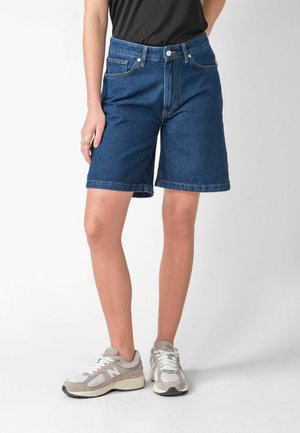 Persoană care poartă pantaloni scurți din denim albastru până la genunchi, top negru și adidași gri New Balance, stând pe un fundal alb.