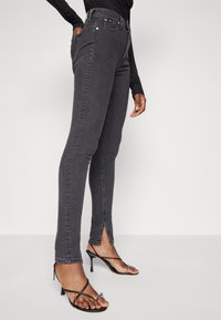 Black high-waisted jeans with a slim fit, featuring front pockets, contrasting stitching, and ankle slits, paired with strappy black heels.