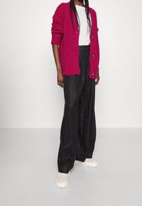 Fuchsiový cardigan s texturovaným pletením, nošený přes bílé tričko, v kombinaci s černými kalhotami s širokými nohavicemi a chunky krémovými botami.