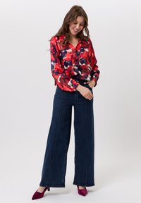 Rode en marineblauwe blouse met patroon, met een kraag, lange mouwen en elastische manchetten, gecombineerd met hoge donkere blauwe wijde jeans en bordeauxrode hakken.