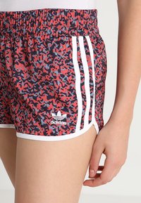 Červené a námořnické křivé sportovní šortky s bílými pruhy, elastickým pasem a malým bílým logem Adidas na přední straně.