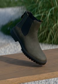 Grünes Leder-Ankle-Boot mit schwarzen elastischen Seitenpaneelen, robustem Profil, schlankem Design und minimalen Nähdetails, präsentiert auf einer Holzoberfläche.