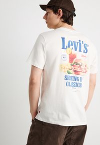 Νέος άνδρας που φοράει λευκό t-shirt Levi's με γραφικό μπέργκερ, πατάτες, ποτό και το κείμενο "Serving Up Classics!" σε μπλε, συνδυασμένο με καφέ παντελόνια και καπέλο.
