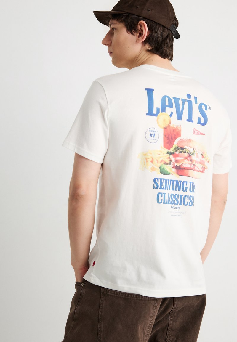 Νέος άνδρας που φοράει λευκό t-shirt Levi's με γραφικό μπέργκερ, πατάτες, ποτό και το κείμενο "Serving Up Classics!" σε μπλε, συνδυασμένο με καφέ παντελόνια και καπέλο.