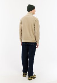 Beige fleece jack met hoge kraag, gecombineerd met donkerblauwe jeans en een groene gebreide beanie; van achteraf weergegeven, met casual bruine schoenen.
