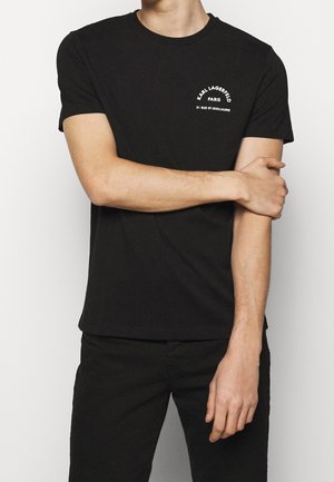 Person trägt ein schwarzes Karl Lagerfeld T-Shirt mit weißem Logo auf der linken Brustseite, schwarze Hose, steht vor einem schlichten Hintergrund.