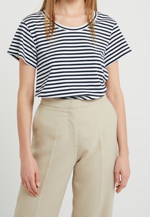 Frau trägt ein kurzärmeliges Shirt mit marineblauen und weißen horizontalen Streifen, das in hoch taillierte beige Hosen gesteckt ist, vor einem schlichten Hintergrund.