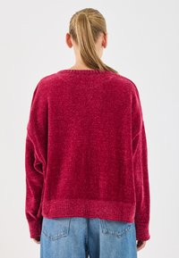 Pull en maille cramoisi avec une coupe décontractée, épaules tombantes et ourlet côtelé. La vue de dos met en avant un tissu texturé. Associé à un jean bleu.