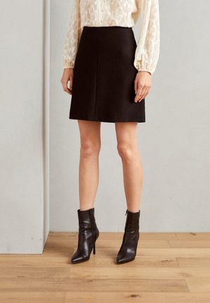 Femme portant une jupe noire mi-longue, un chemisier crème à manches longues avec des détails de boutons, et des bottines noires à talons hauts.