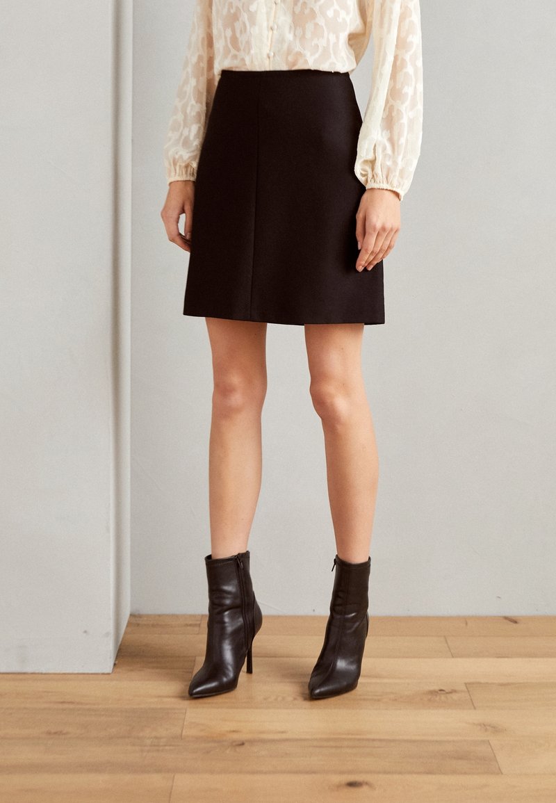 Femme portant une jupe noire mi-longue, un chemisier crème à manches longues avec des détails de boutons, et des bottines noires à talons hauts.