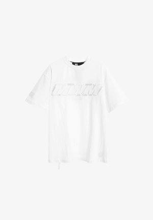 Camiseta blanca de algodón con mangas cortas, que cuenta con un logo en relieve en gris claro en el pecho y una etiqueta en el lateral. Textura suave.