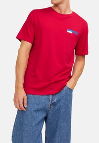 Rotes Baumwoll-T-Shirt mit Rundhalsausschnitt und kurzen Ärmeln, mit einem blau-weißen grafischen Akzent im linken Brustbereich.