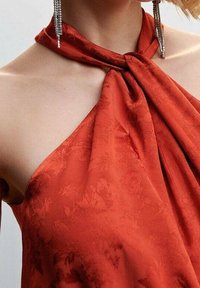 Top halter de satén rojo con un nudo en el escote, que presenta un sutil patrón floral y una textura suave. Pendientes de gota plateados visibles.