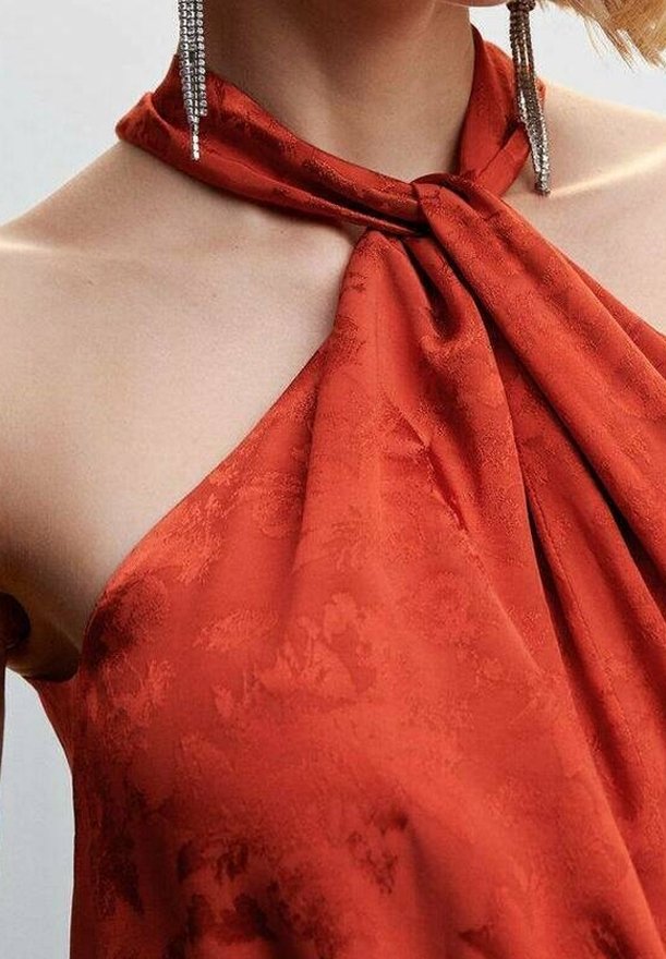 Top halter de satén rojo con un nudo en el escote, que presenta un sutil patrón floral y una textura suave. Pendientes de gota plateados visibles.