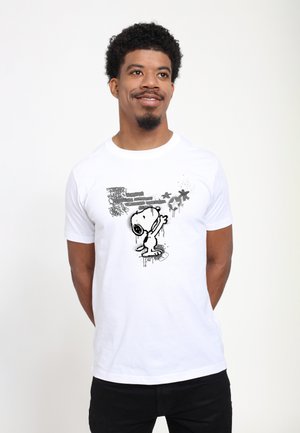 Homme aux cheveux bouclés souriant, portant un t-shirt blanc montrant Snoopy tendant la main vers trois étoiles et des formes noires abstraites.