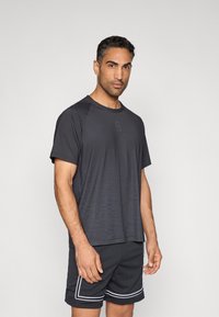 Αθλητική μπλούζα σκούρου γκρι με διακριτικό ριγέ μοτίβο, συνδυασμένη με μαύρο σορτς από δίχτυ με λευκές λεπτομέρειες και χαλαρή εφαρμογή.