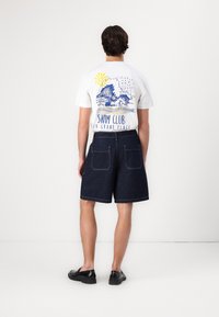 Hvit T-skjorte med blå og gul grafisk design, kombinert med mørke jeansshorts. Shortsen har to baklommer og en rett passform.