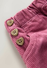 Tessuto in velluto a coste rosa con tre bottoni marroni a forma di cuore sul lato sinistro. Superficie testurizzata con righe verticali e cucitura sul bordo.