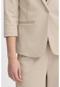 Nahaufnahme einer Person in einem beigefarbenen maßgeschneiderten Anzug mit einreihigem Sakko und passenden Hosen, der Arm entspannt an der Seite.