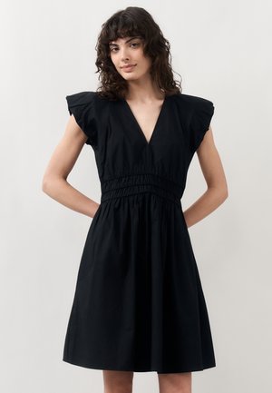 Woman with curly brown hair wearing a black sleeveless dress with a deep V-neck and gathered waist, standing with hands behind her back.