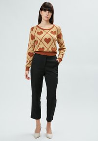 Beige Pullover mit braunem Herzmuster in einem Diamantdesign, kombiniert mit schwarzen Hosen und beige spitzen Pumps. Weiche Textur sichtbar.