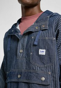 Denim-Pullover mit hellblauen vertikalen Streifen, Kapuze und Fronttaschen. Verfügt über silberne Druckknöpfe und ein Lee-Logo auf der Brusttasche.