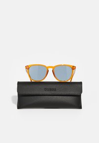Orange solglasögon med blå linser placerade ovanför ett svart läderskydd med den präglade "GUESS"-logotypen och lockstängning.