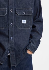 Camisa vaquera en azul oscuro con dos bolsillos en el pecho, cierre de botones y parche con el logo. Presenta un corte relajado y detalles de costuras visibles.