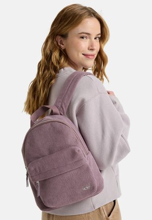 Lila Cordura-Rucksack mit einer vorderen Reißverschlusstasche, gepolsterten Schulterriemen und Logo-Detail. Getragen von einer Person in einem leichten Pullover.