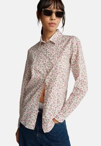 Camicia bianca con bottoni, motivo floreale in piccoli dettagli rosa e verdi, maniche lunghe e una leggera apertura laterale. Abbinata a jeans scuri.