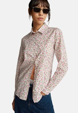 Witte blouse met een klein roze en groene bloemenpatroon, lange mouwen en een minimale zijsplit. Gecombineerd met donkere denim jeans.