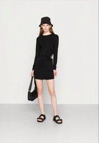 Vestido preto de manga comprida com franzidos laterais, design ajustado e detalhe de recorte. Combinado com sandálias plataforma pretas e um chapéu tipo balde a condizer.