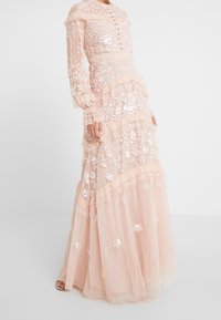Lång, mjuk persikofärgad klänning med spetsdetaljer, blommig brodyr och paljetter, som har en hög halsringning, knappstängning och volangskikt.