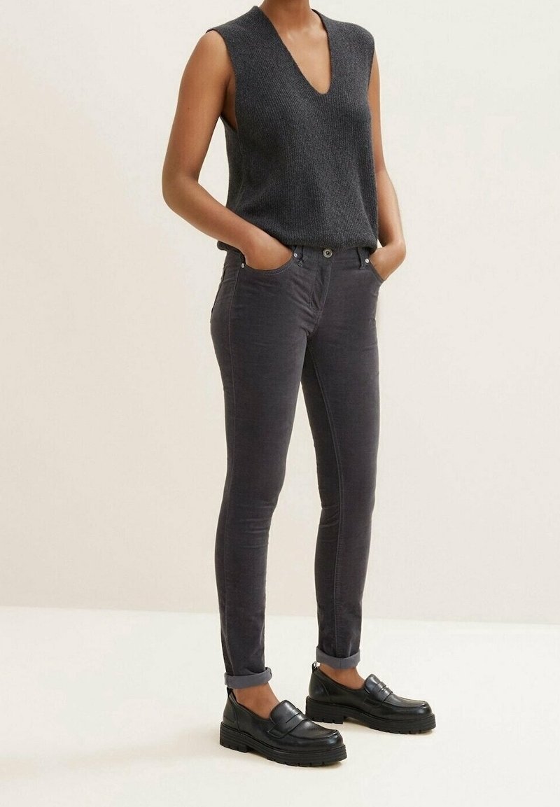 Personne portant un haut sans manches en maille gris foncé, un pantalon gris foncé ajusté retroussé, et des mocassins à plateforme noirs, debout les mains dans les poches.