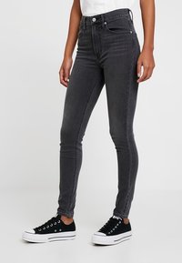 Jeans negros de cintura alta en un corte ajustado, con cinco bolsillos, pasadores para cinturón y costuras en contraste. Combinados con zapatillas negras.