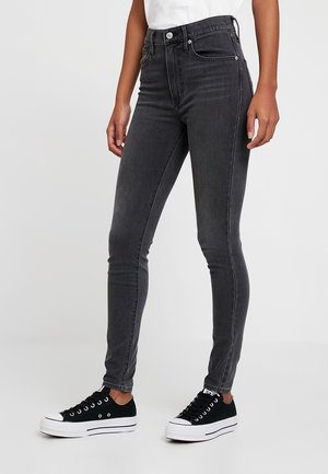 Jeans negros de cintura alta en un corte ajustado, con cinco bolsillos, pasadores para cinturón y costuras en contraste. Combinados con zapatillas negras.