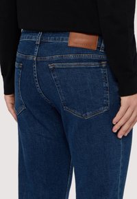 Tmavě modré džínové kalhoty s texturovaným povrchem, s zadními kapsami a hnědou koženou štítkou s nápisem "TRUSSARDI" v pase.