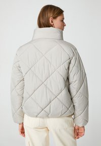 Chaqueta acolchada puffer de color gris claro con cuello alto, que presenta un patrón de diamante y un ajuste relajado. Textura suave y costuras de hombro redondeadas.