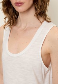 Balta be rankovių palaidinė su apvaliu iškirpimu, pagaminta iš lengvo, tekstūrinio audinio; turi švarius kraštus ir nežymius spalvų skirtumus.