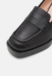 Black leather loafer with a rounded toe and subtle stitching details. Thick rubber sole provides added height and durability. Textured accent band.