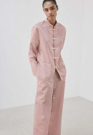 Femme portant une veste à rayures verticales rose et blanche à manches longues et un pantalon large assorti, debout les mains dans les poches.