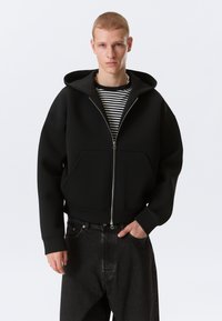 Jeune homme portant un sweat à capuche zippé noir par-dessus une chemise rayée noir et blanc et un jean foncé, se tenant devant un fond clair uni.