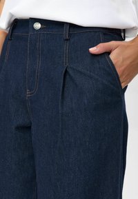 Close-up of dark blue denim pants with orange stitching, front pleat, metal button, and a hand resting in the right pocket.