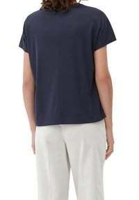 Tummansininen lyhythihainen paita pehmeästä, tekstuurilla varustetusta kankaasta, rento malli ja takasauman yksityiskohta, yhdistettynä kermanvärisiin housuihin.