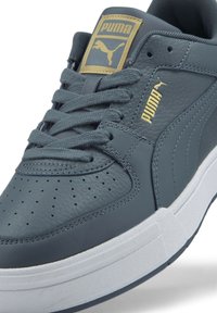 Baskets en cuir grises avec un embout perforé, des lacets plats et un logo doré Puma sur la languette. Semelle en caoutchouc blanche avec une texture antidérapante.