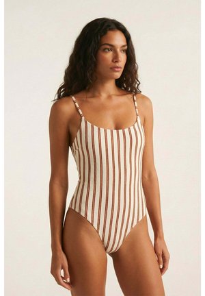 Mujer con cabello rizado largo y oscuro, vestida con un bañador blanco de una pieza con finas rayas verticales marrones, de pie frente a un fondo claro y liso.